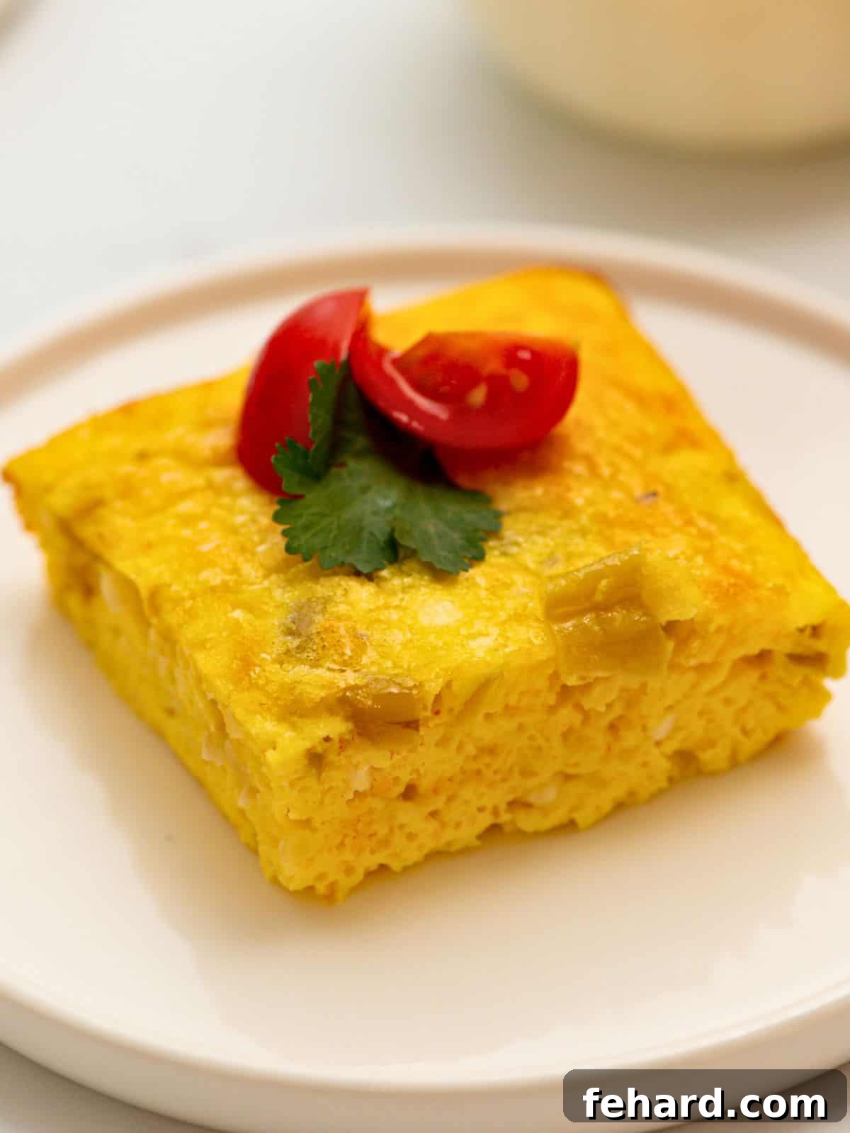 A perfectly portioned slice of fluffy Green Chile Egg Casserole, adorned with fresh diced tomatoes and vibrant cilantro, rests on a crisp white plate, highlighting its rich, cheesy layers and mild green chile flecks.