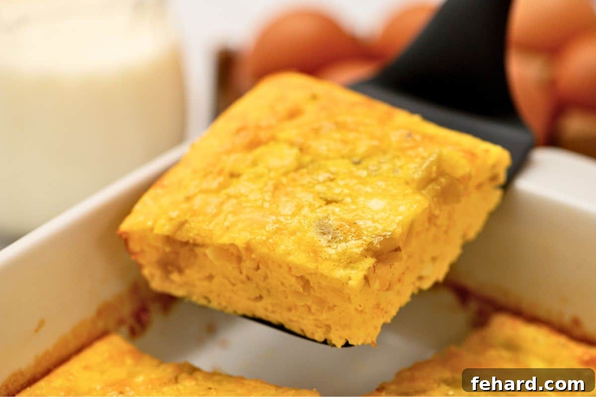 A serving spoon expertly lifts a creamy, cheesy slice of egg casserole, revealing the cottage cheese and green chiles, from a pristine white casserole dish, ready to be enjoyed.