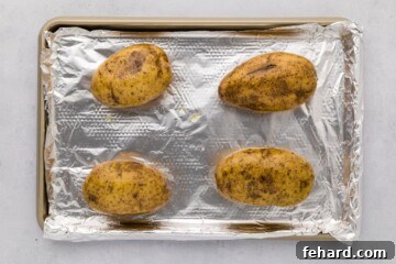 Four russet potatoes on a baking sheet lined with foil, ready for baking.
