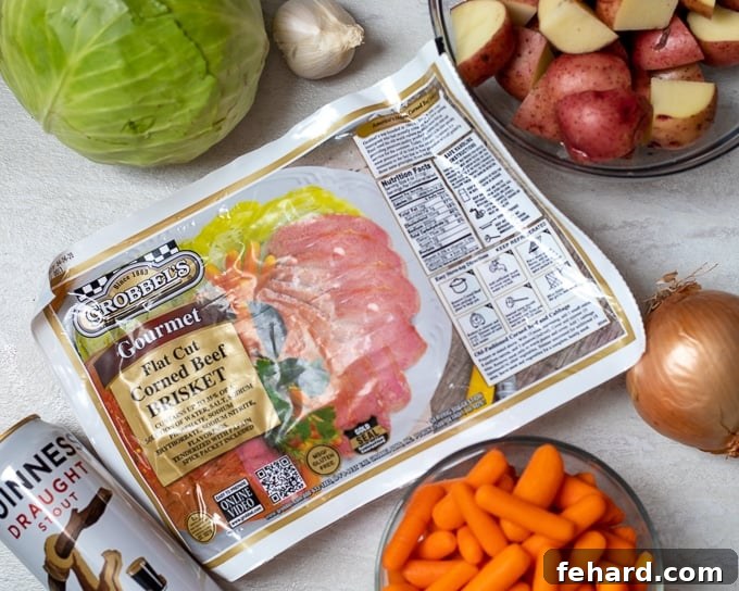 Ingredients for corned beef and cabbage laid out on a white cutting board: corned beef brisket, green cabbage, yellow onion, red potatoes, carrots, and a bottle of stout beer.