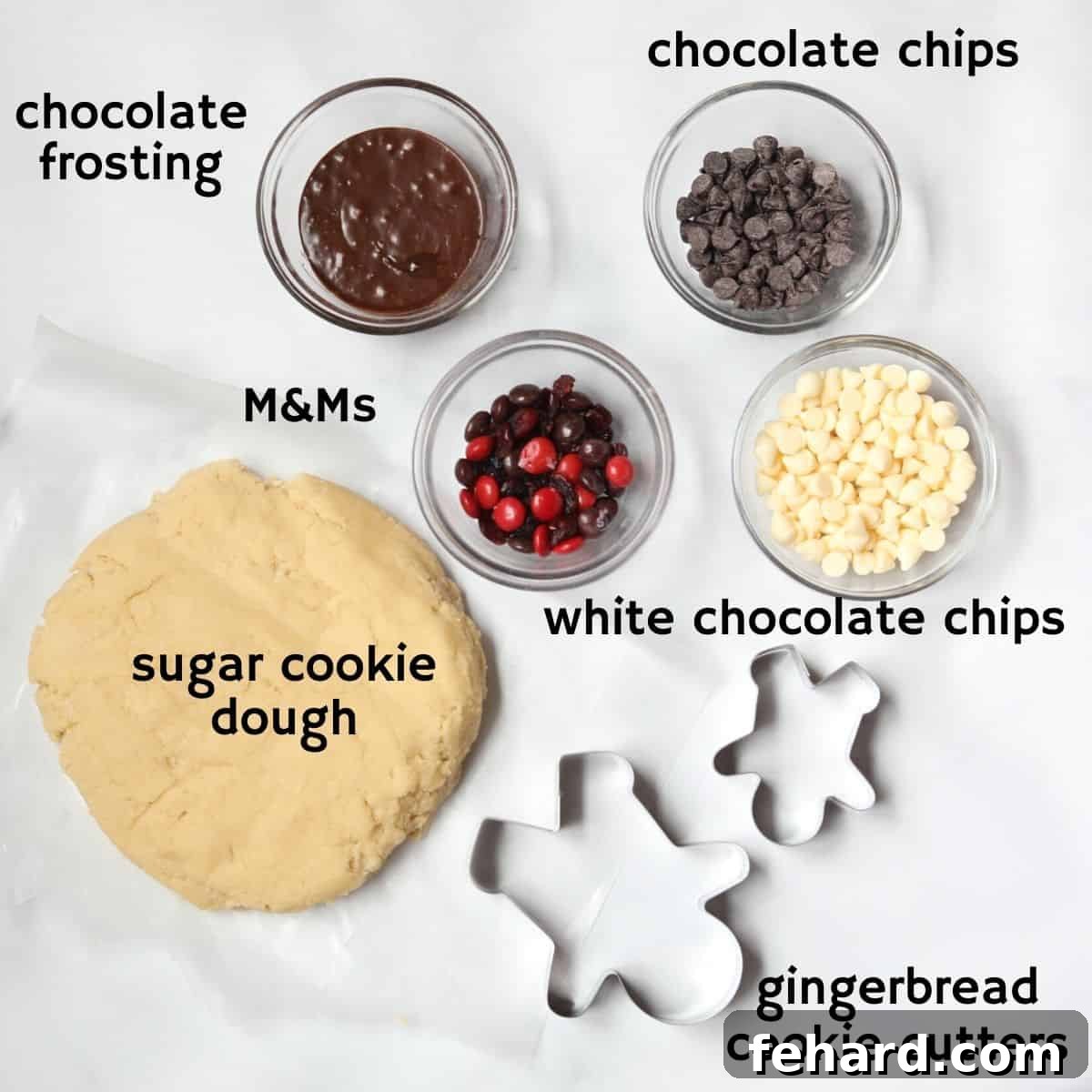 Arrangement of ingredients for making reindeer cookies, including sugar cookie dough, chocolate frosting, candy eyes, M&Ms, and melted chocolate, on a clean white kitchen counter.