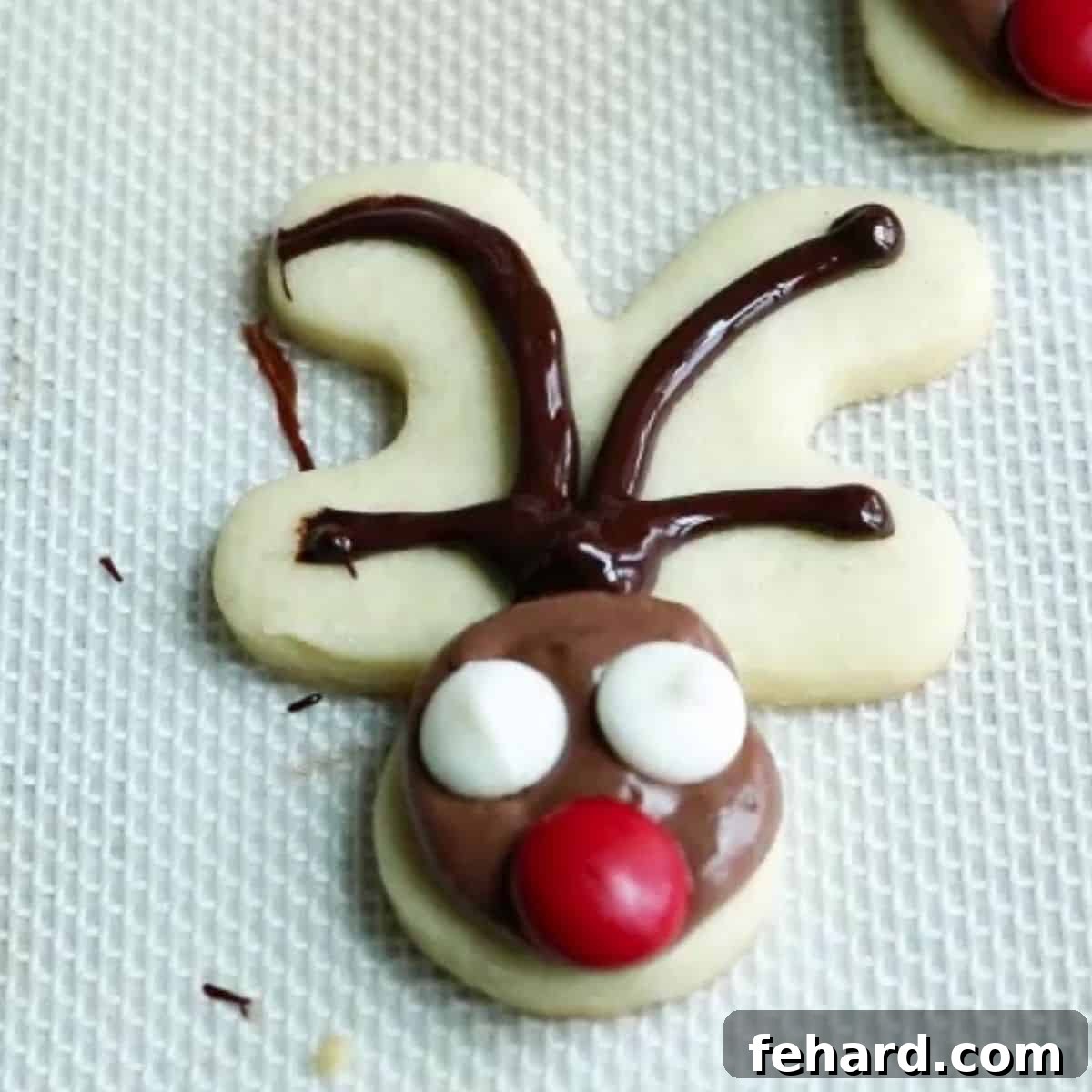 A gingerbread man cookie decorated as a reindeer, with chocolate frosting, a candy nose, and white chocolate chips for eyes, awaiting its antlers and pupils.
