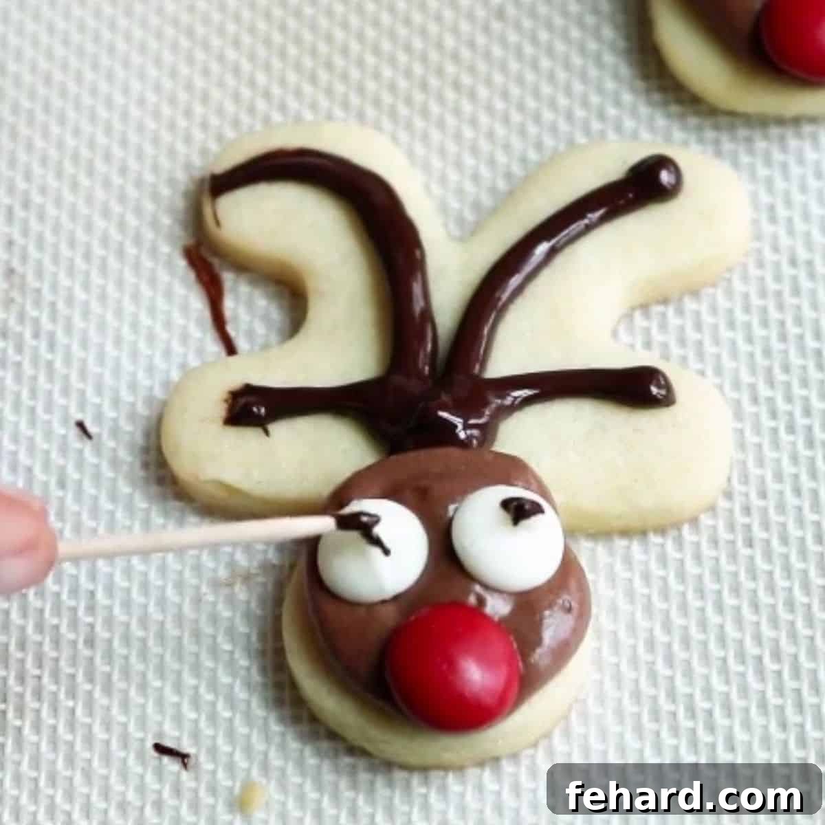A close-up shot of a reindeer cookie, showing a toothpick being used to add a small dot of melted chocolate to a white chocolate chip eye, creating a pupil.