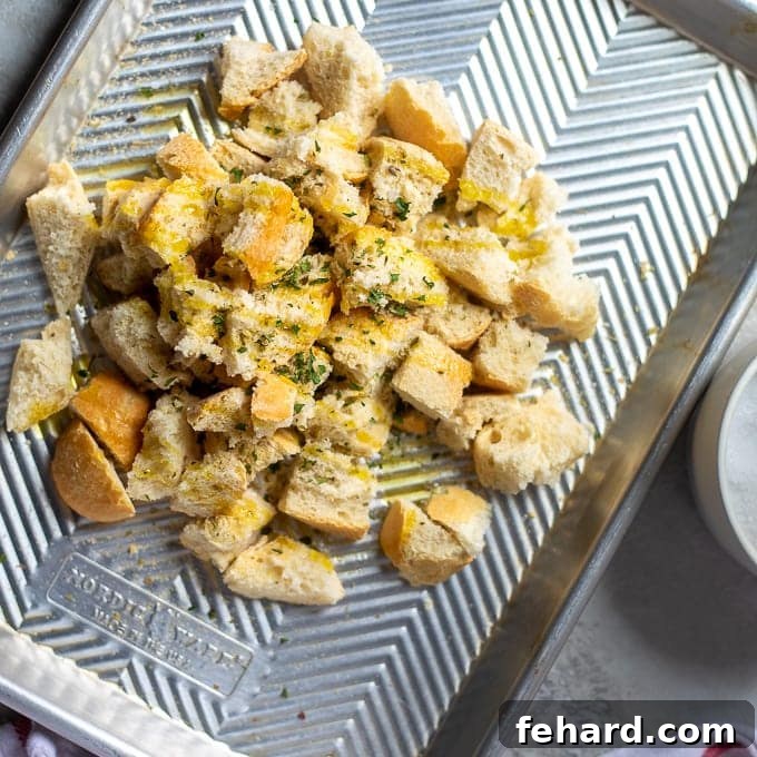 Close-up of bread cubes on a baking sheet, glistening with olive oil and sprinkled with a blend of dried herbs and spices, ready for tossing.