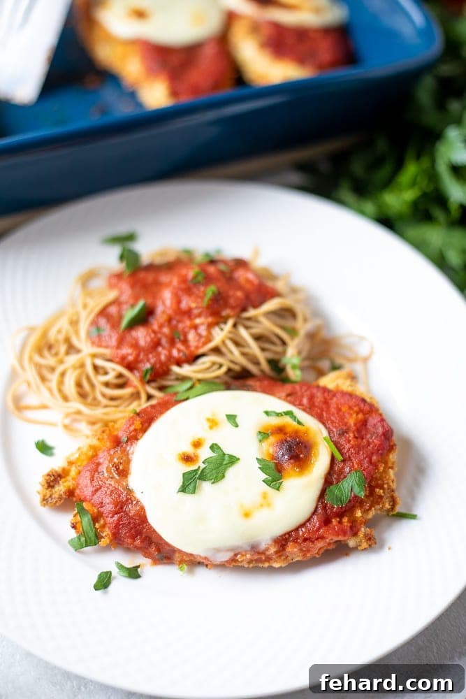 Crispy Baked Chicken Parmesan on a white plate next to a serving of spaghetti and fresh basil.