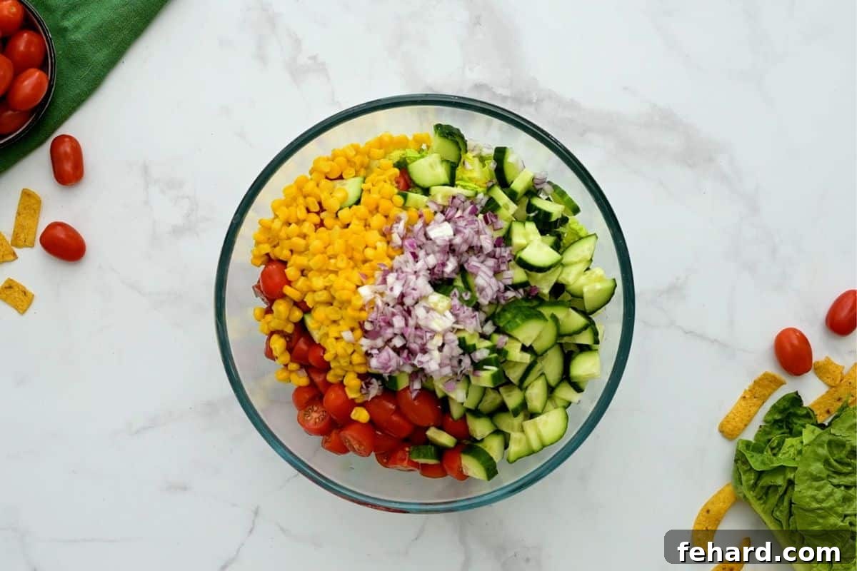 A large bowl filled with chopped lettuce, diced cucumbers, minced red onions, sliced tomatoes, and corn kernels, ready to be tossed.