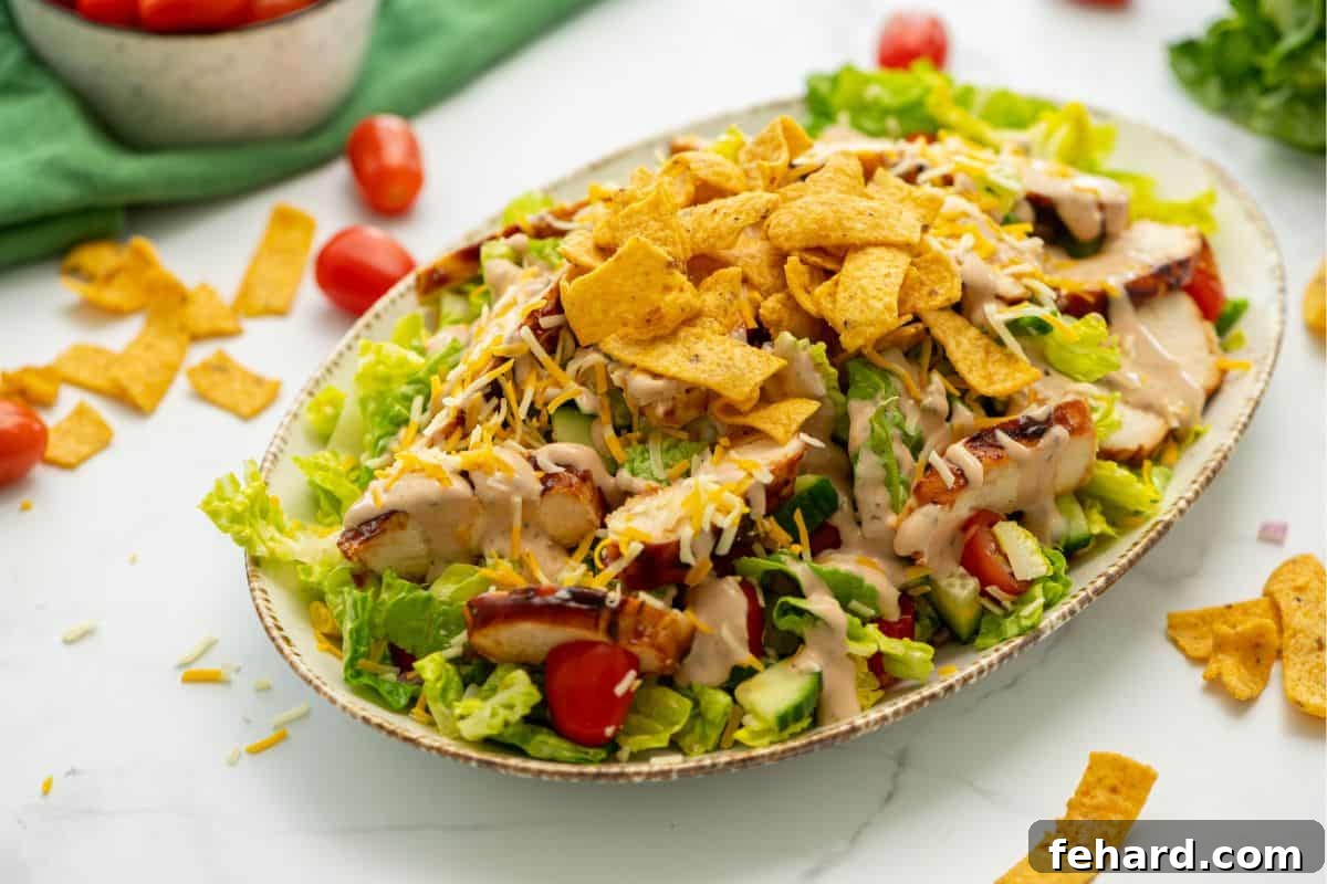 An assembled BBQ Chicken Salad with sliced chicken, shredded cheese, corn chips, and a drizzle of BBQ Ranch Dressing, with tomatoes in the background.