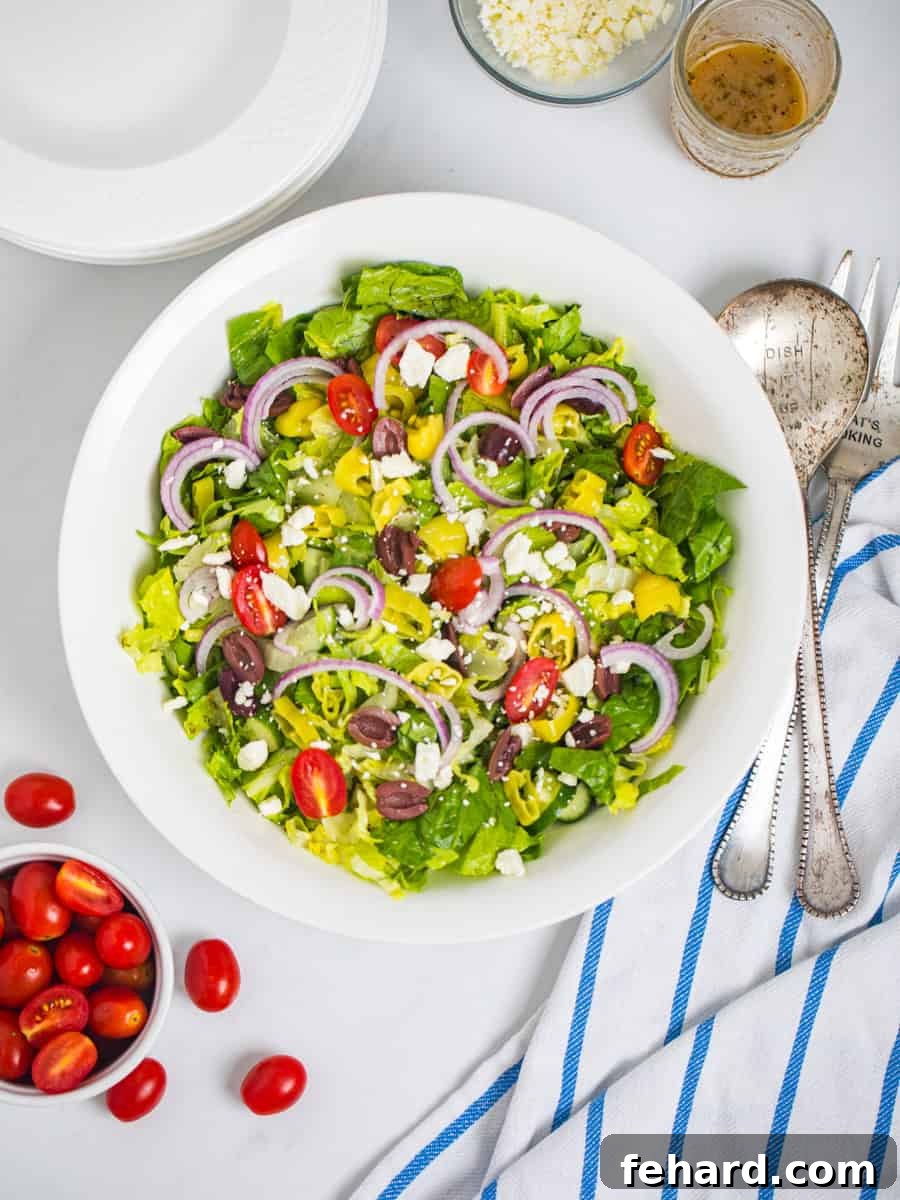 Copycat Panera Greek Salad Recipe A large white bowl brimming with freshly tossed Greek salad, showcasing crisp lettuce, colorful grape tomatoes, kalamata olives, red onions, cucumbers, and crumbled feta cheese, with serving spoons nestled beside a handful of fresh grape tomatoes.