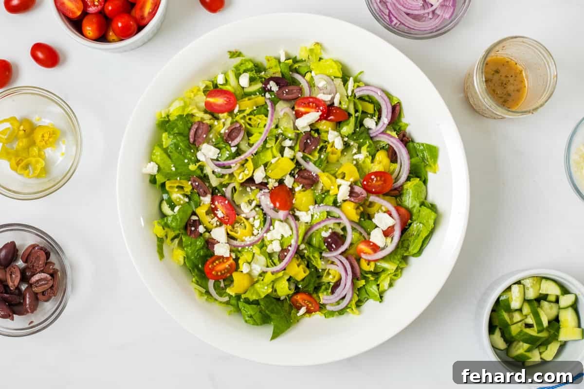 Tossed Greek Salad A beautifully presented tossed Greek salad in a large white bowl, featuring crisp lettuce, bright red onions, sliced pepperoncini peppers, fresh cucumbers, kalamata olives, and crumbled feta cheese, ready to be served. This image highlights the delicious combination of flavors and textures in the homemade Panera Greek Salad.