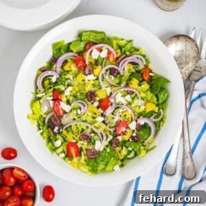 Tossed greek salad with Greek vinaigrette in large white bowl next to grape tomatoes and serving spoons.
