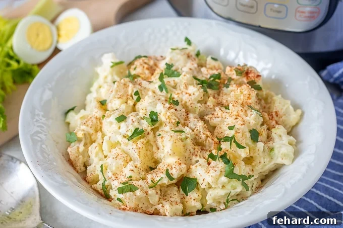 Instant Pot Potato Salad Ready to Serve A pristine white bowl brimming with freshly made Instant Pot Potato Salad, garnished with a sprinkle of paprika and fresh parsley. Hard-boiled eggs and celery are visible in the background, hinting at the key ingredients.