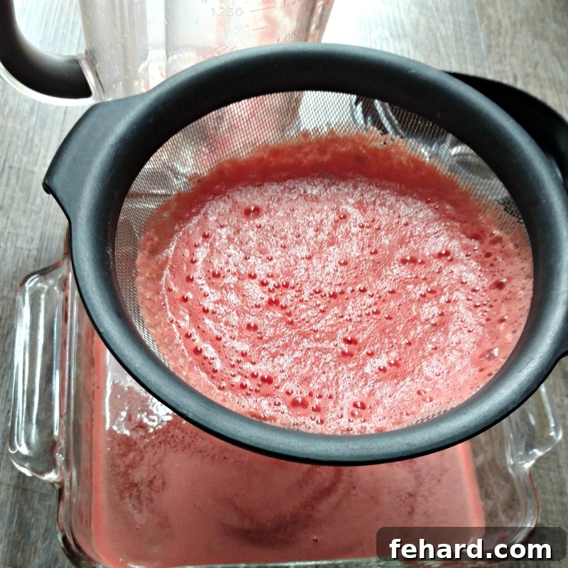 Watermelon juice being strained through a fine mesh strainer into a glass baking dish, ensuring a smooth granita.