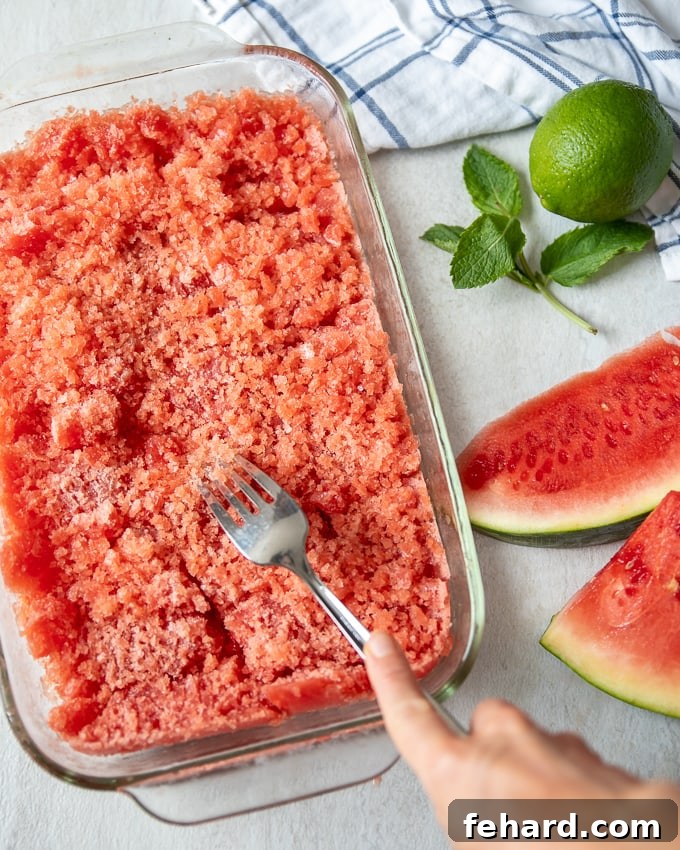 Close-up of a fork scraping the semi-frozen watermelon granita mixture in a clear glass baking dish, forming ice crystals.