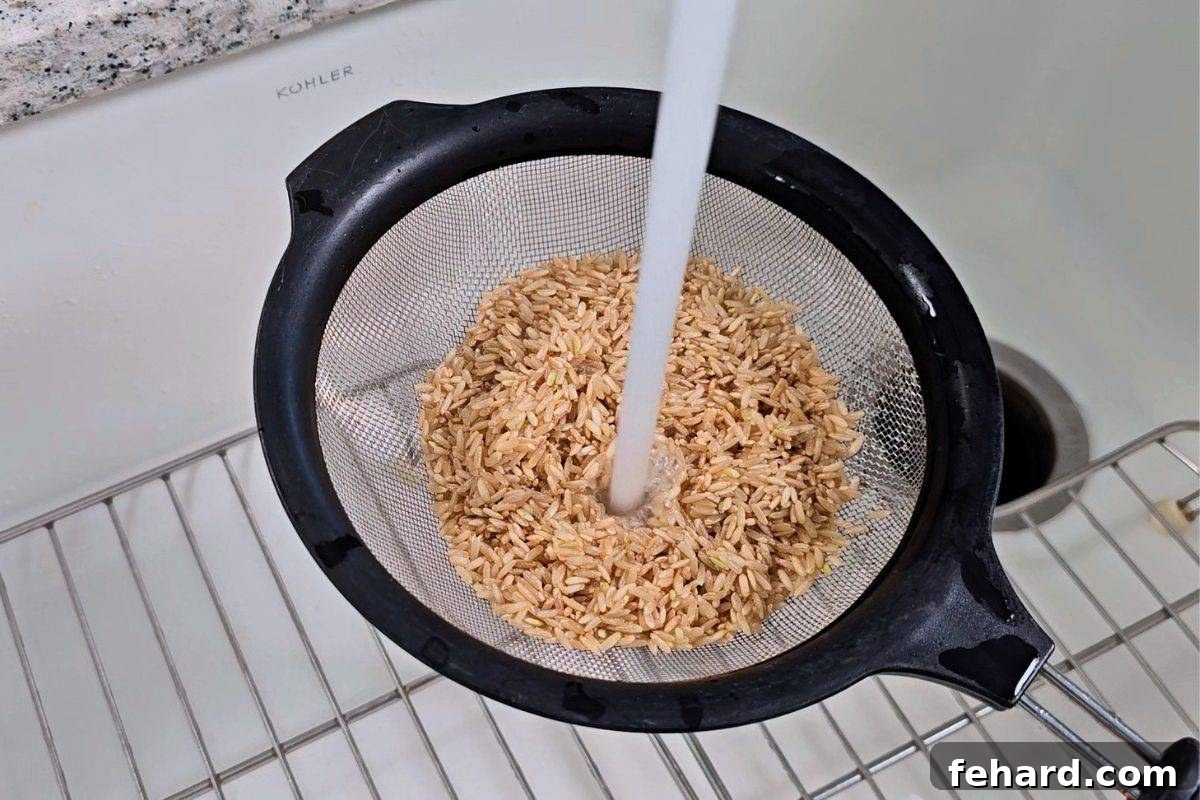 Brown rice inside a fine mesh strainer with cold tap water running over it, showing the rinsing process.