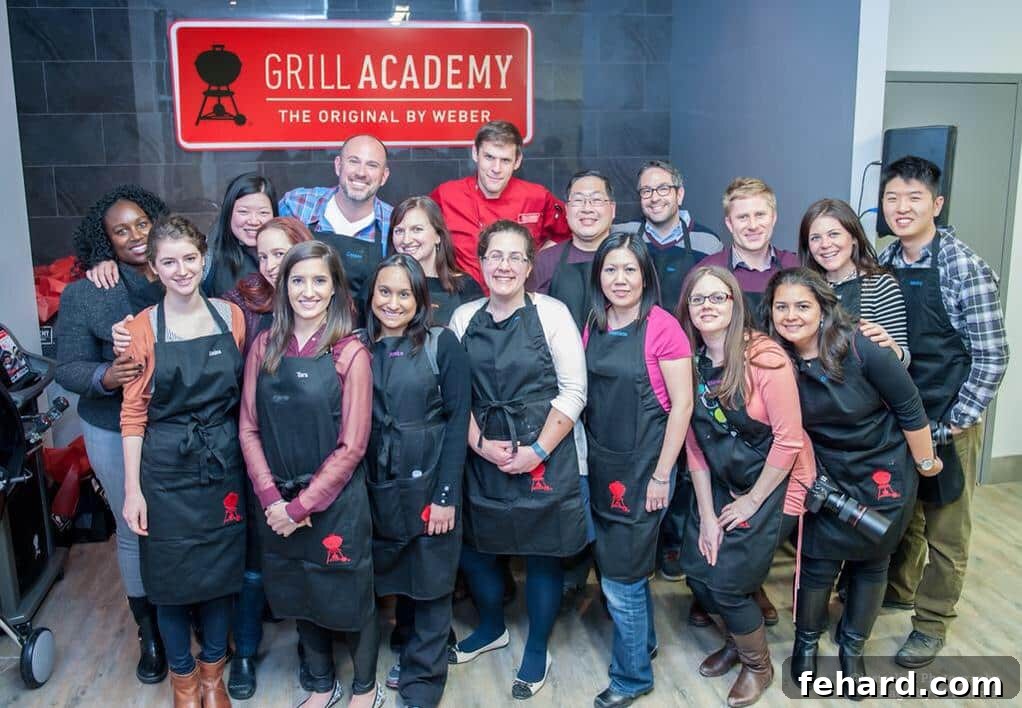 A group of smiling people proudly wearing black Weber aprons, happily gathered for a group photo in front of the iconic red Grill Academy sign, celebrating their shared grilling experience and camaraderie.