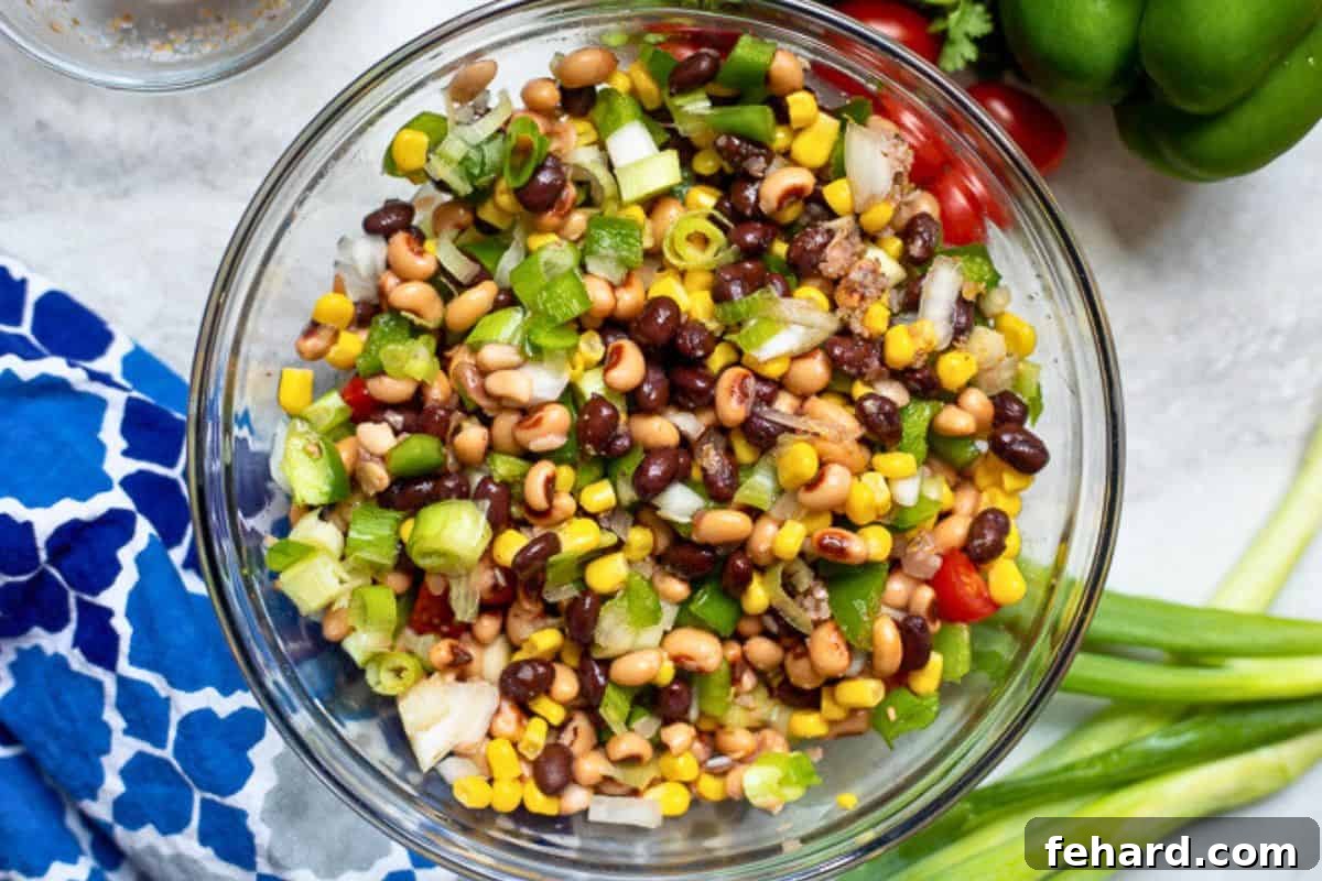 Recipe Cowboy Caviar Mix A vibrant mix of black-eyed peas, black beans, corn, green onions, bell peppers, and tomatoes are tossed in a tangy Italian-style dressing within a large mixing bowl, showcasing the colorful medley of ingredients.