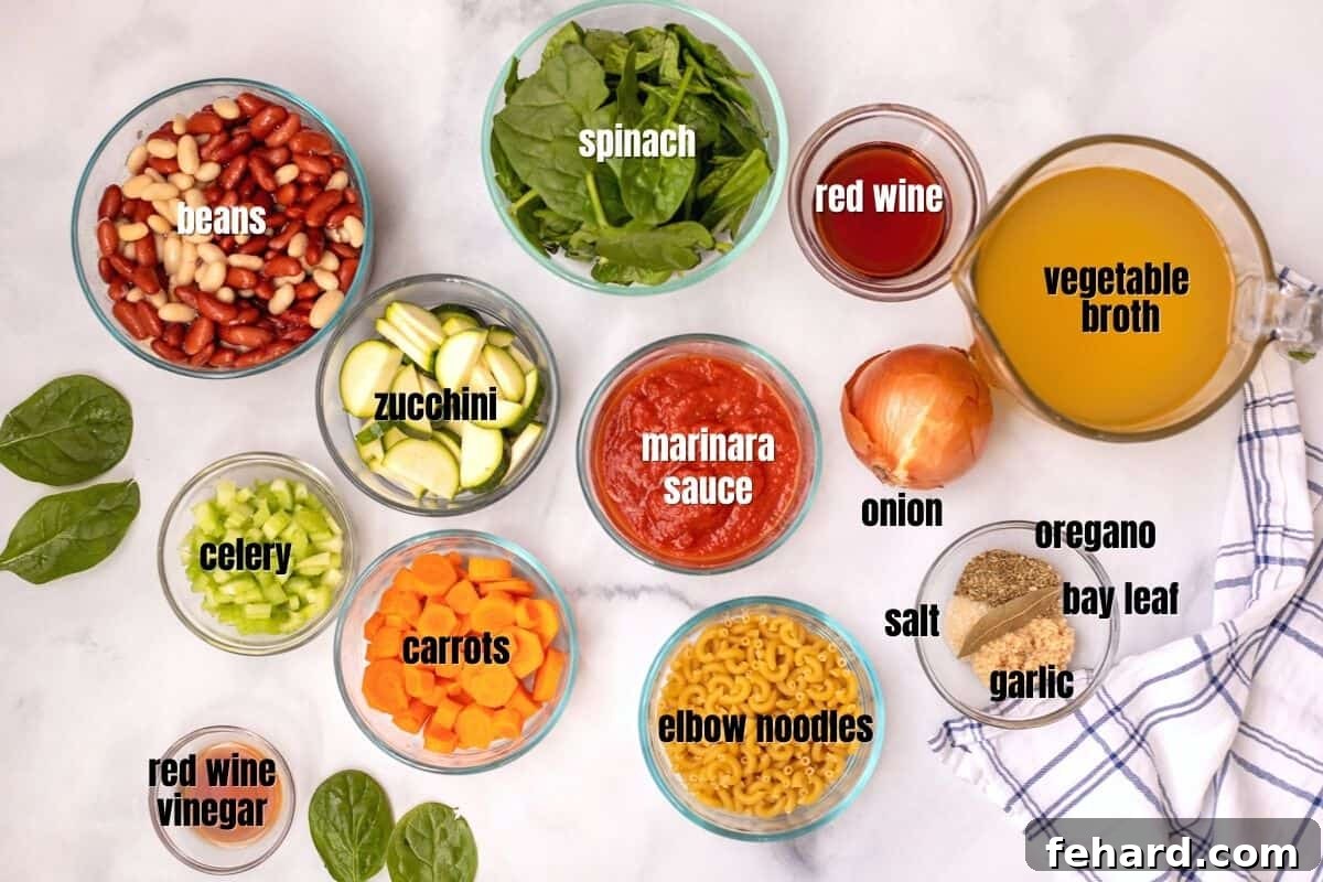 Fresh ingredients laid out on a white countertop, including onions, carrots, celery, garlic, zucchini, canned beans, pasta, marinara, and herbs, all ready for making minestrone.