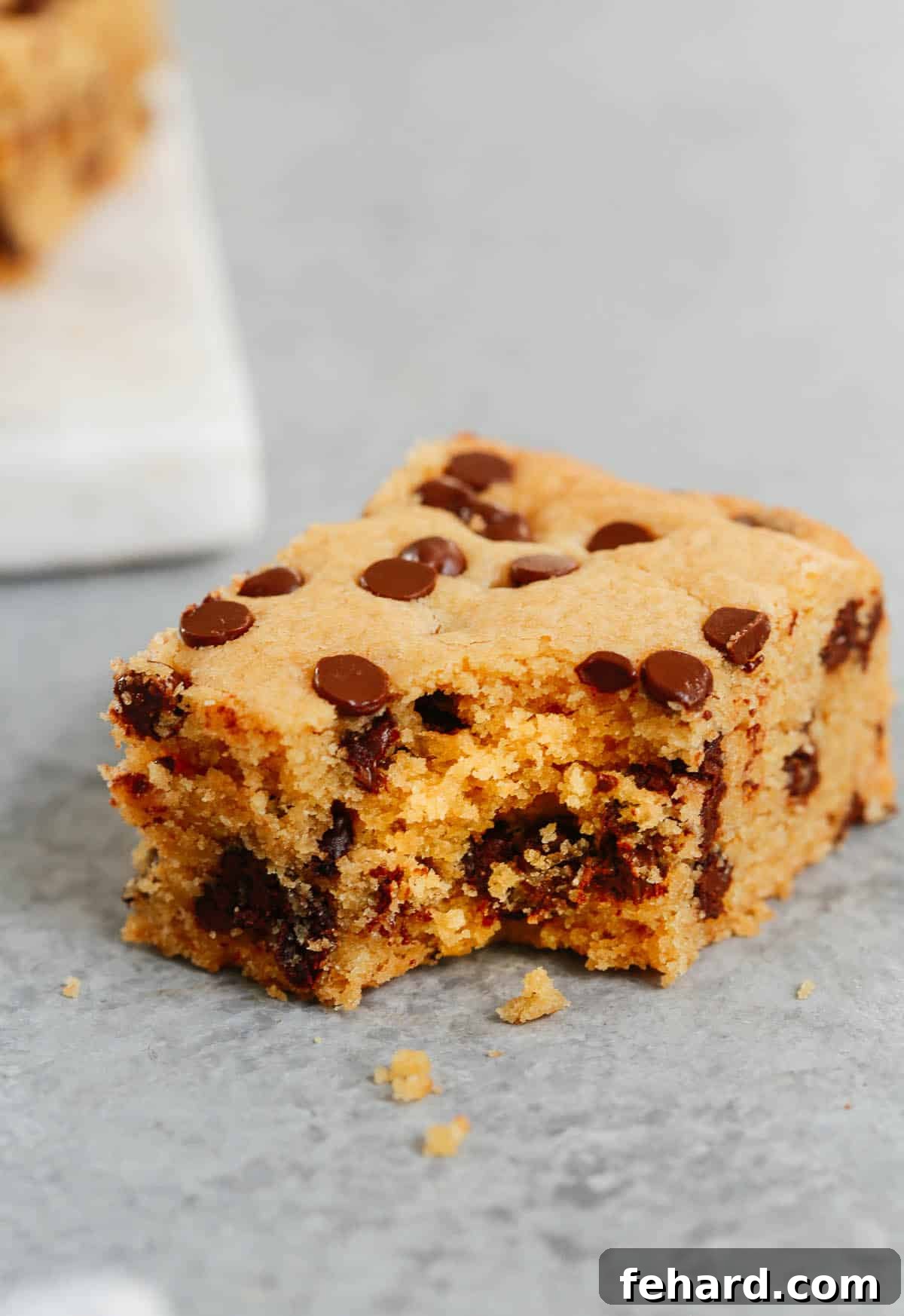 Close up of a chocolate chip cookie bar with a bite taken out, showing its soft, moist interior and abundance of chocolate.