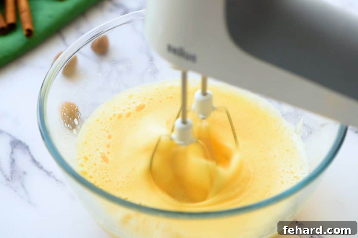 How to Whip Eggs for Pumpkin Cake Roll A handheld mixer vigorously beating eggs in a clear mixing bowl, transforming them into a light, airy, and fluffy consistency.