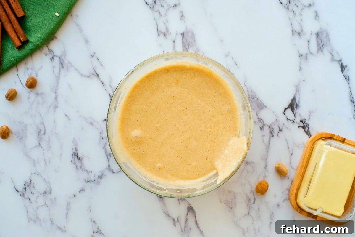 Pumpkin Roll Cake Batter Ready to Bake A large mixing bowl filled with light orange pumpkin sponge cake batter, with a spatula gently folding in the dry ingredients.