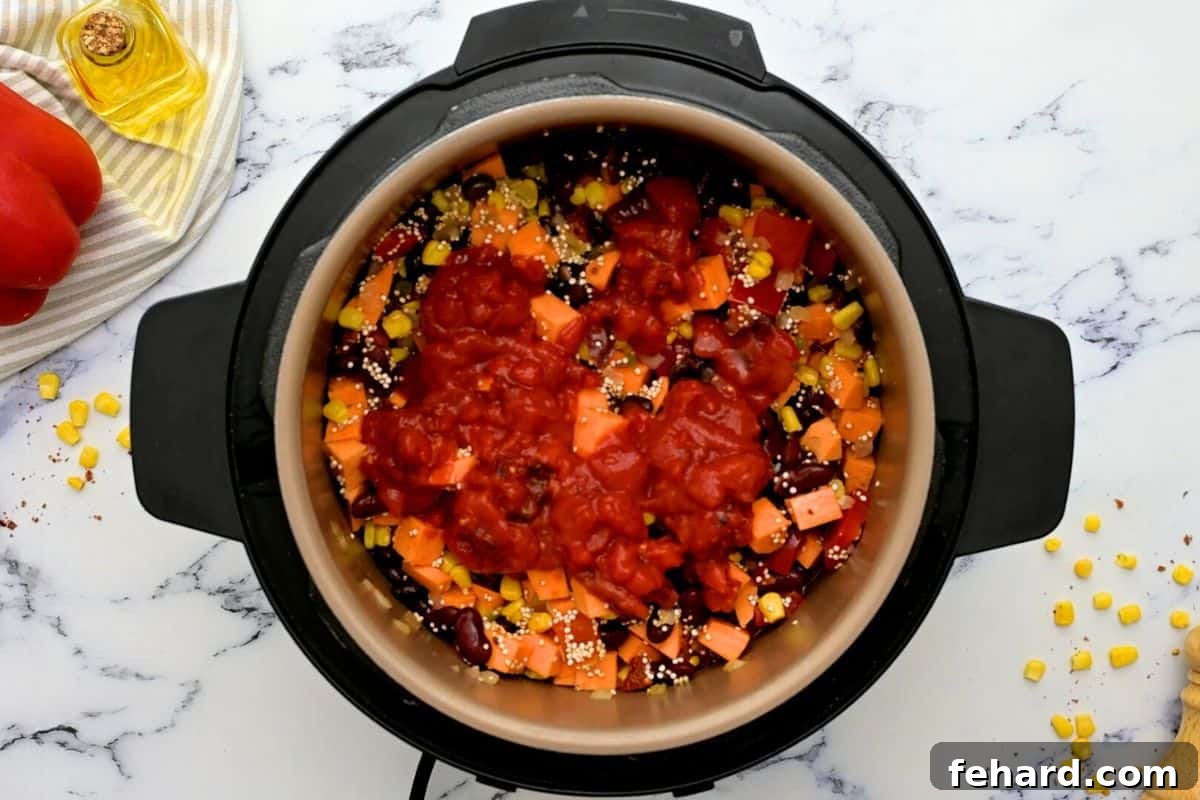 Various chili ingredients, including sweet potatoes, black beans, quinoa, and corn, carefully layered inside the Instant Pot. A vibrant top layer of diced tomatoes sits unstirred, ready for pressure cooking.