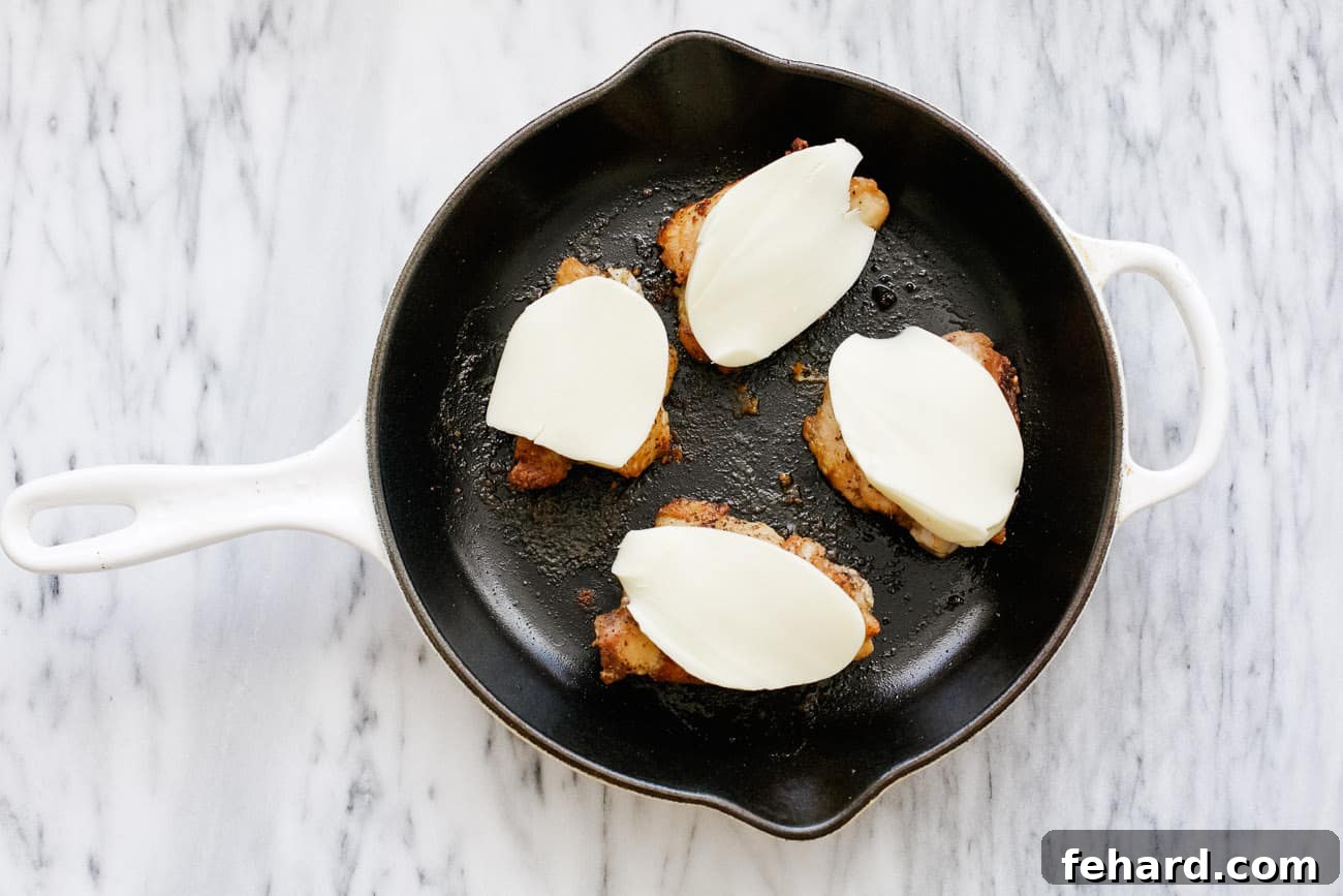 Melted mozzarella cheese generously layered on top of baked chicken thighs in a skillet, showcasing its gooey texture.
