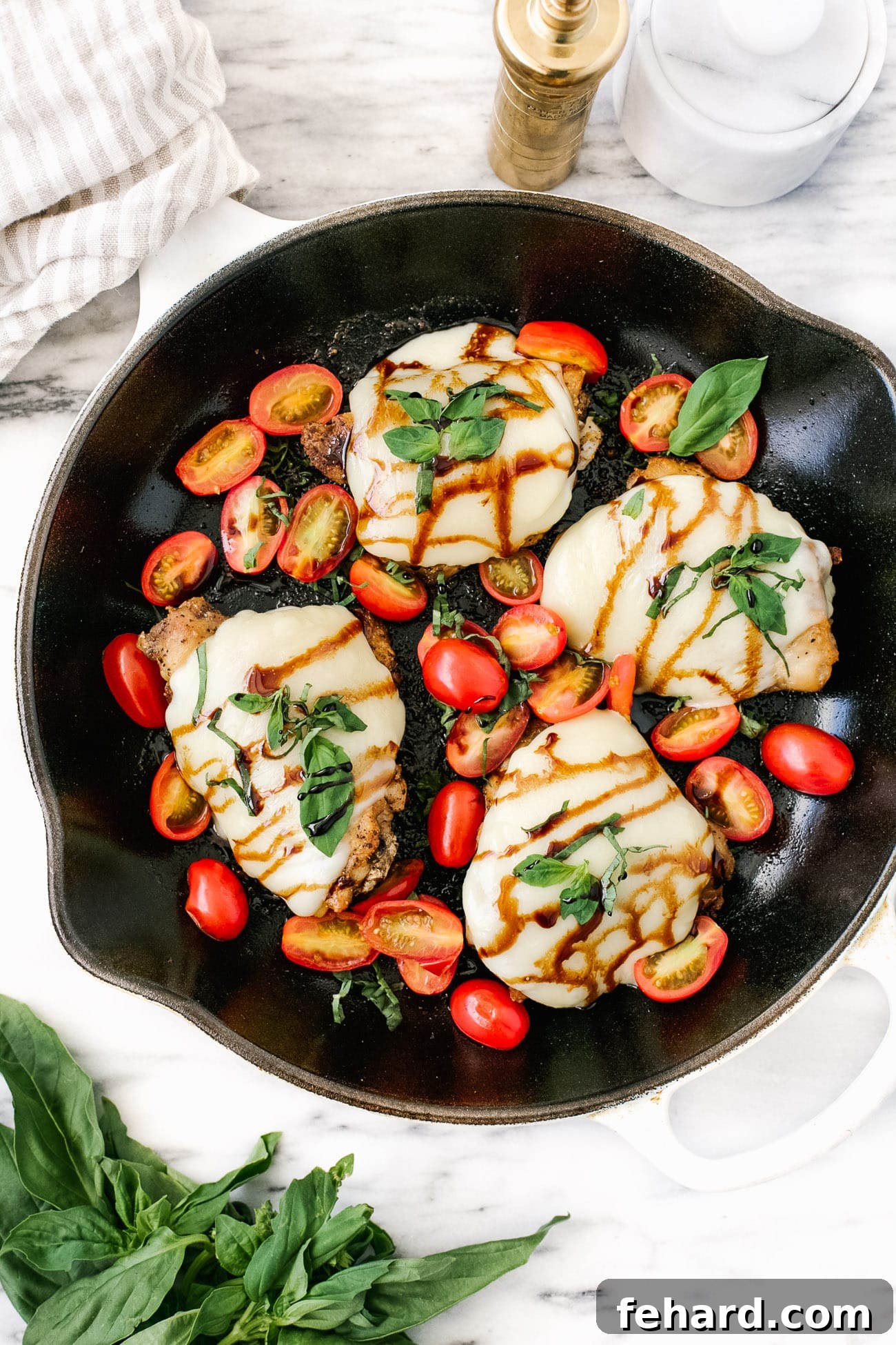 An overhead view of a cast iron skillet filled with baked caprese chicken thighs, garnished with fresh basil and cherry tomatoes.