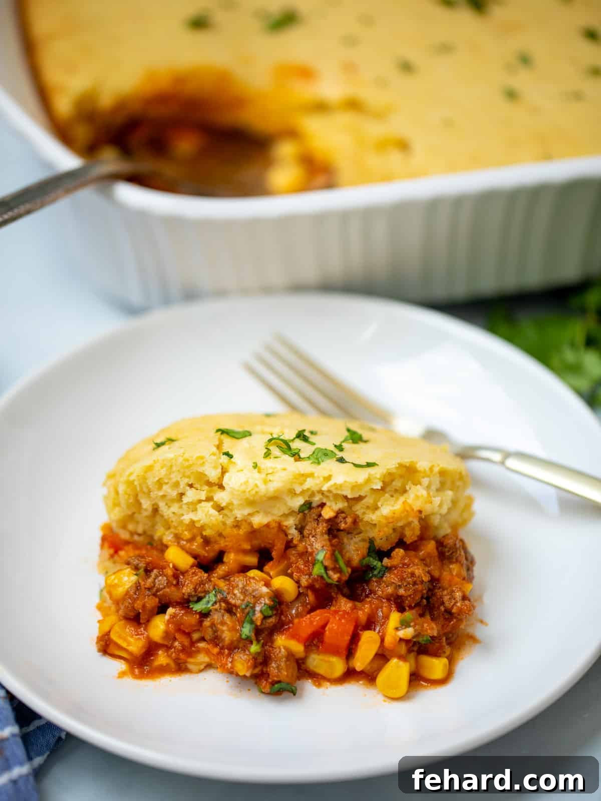 A delicious slice of golden-topped Tamale Casserole, also known as Tamale Pie, served on a white plate with the full casserole dish blurred in the background, showcasing its comforting appeal and vibrant flavors.