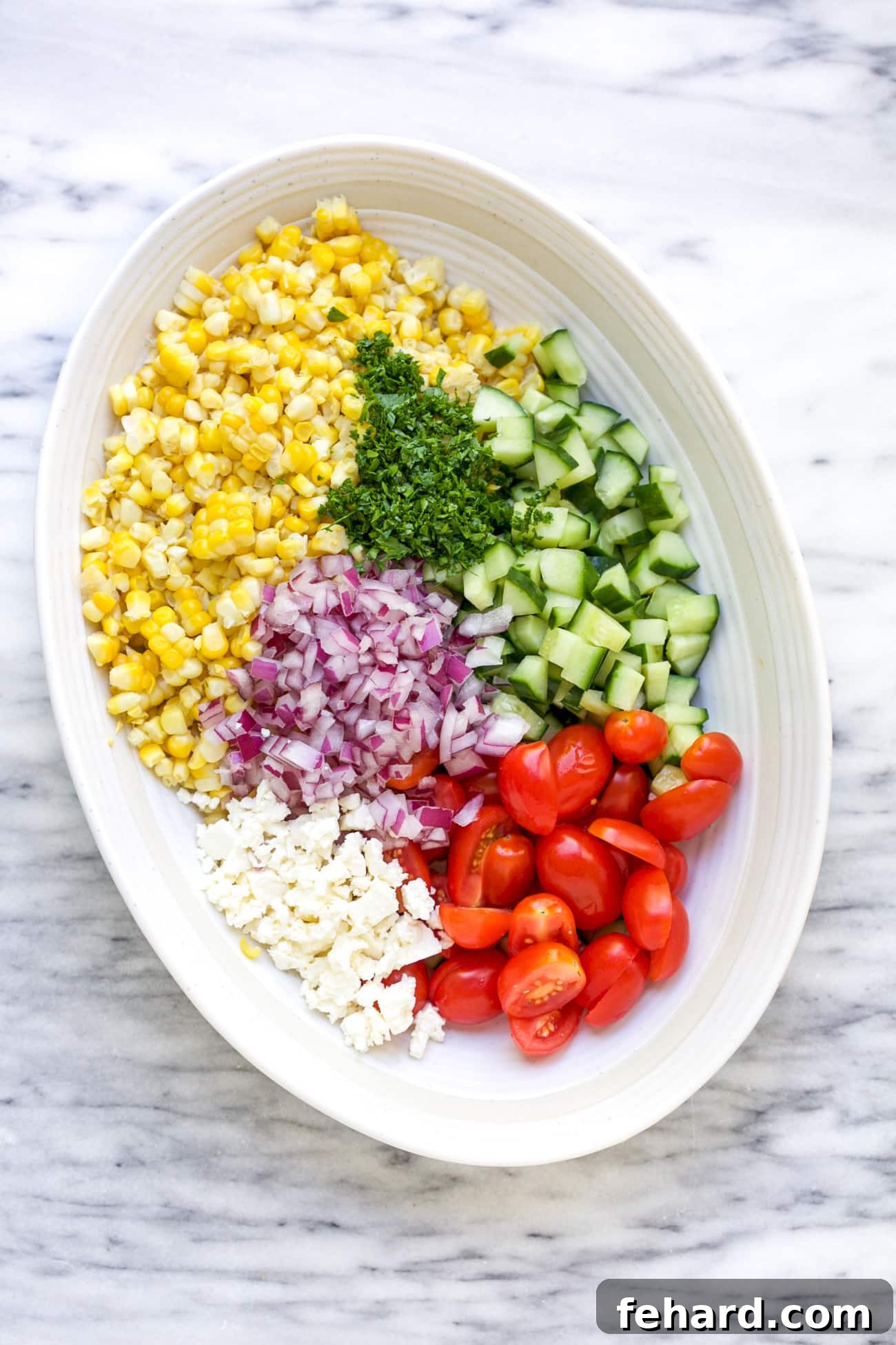 Zesty Corn Salad 5 Prepared ingredients for corn salad, including cooked corn kernels, halved cherry tomatoes, diced cucumber, red onion, and crumbled feta, in a large white mixing bowl on a marble surface.