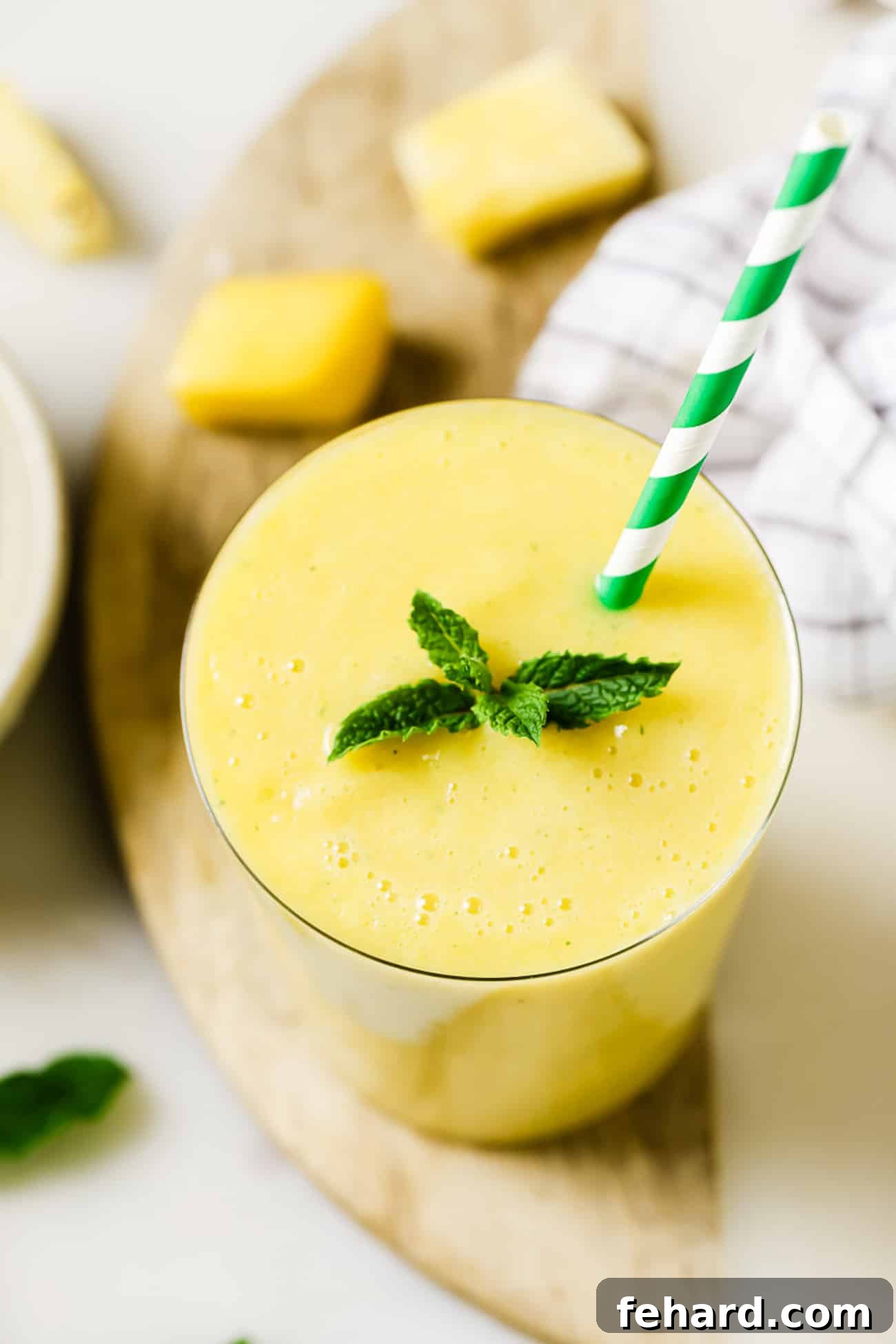 A tall glass of pineapple mango smoothie, garnished with a sprig of fresh mint, on a light background.