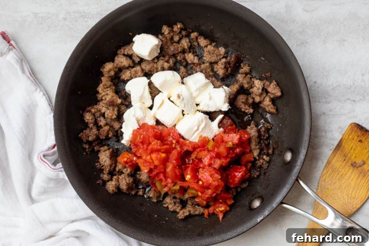 Browned sausage in skillet with undrained Rotel tomatoes and cubed cream cheese, ready to be melted together.