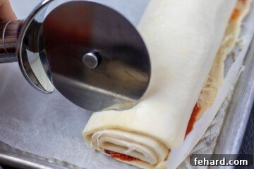 Pizza wheel cutting a roll of puff pastry that is filled with pizza toppings. The dough is being sliced into individual pinwheels.