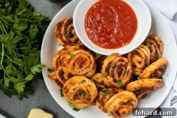 Puff pastry pizza roll-ups in white serving dish next to pizza sauce. The finished, golden-brown roll-ups are presented in a dish.