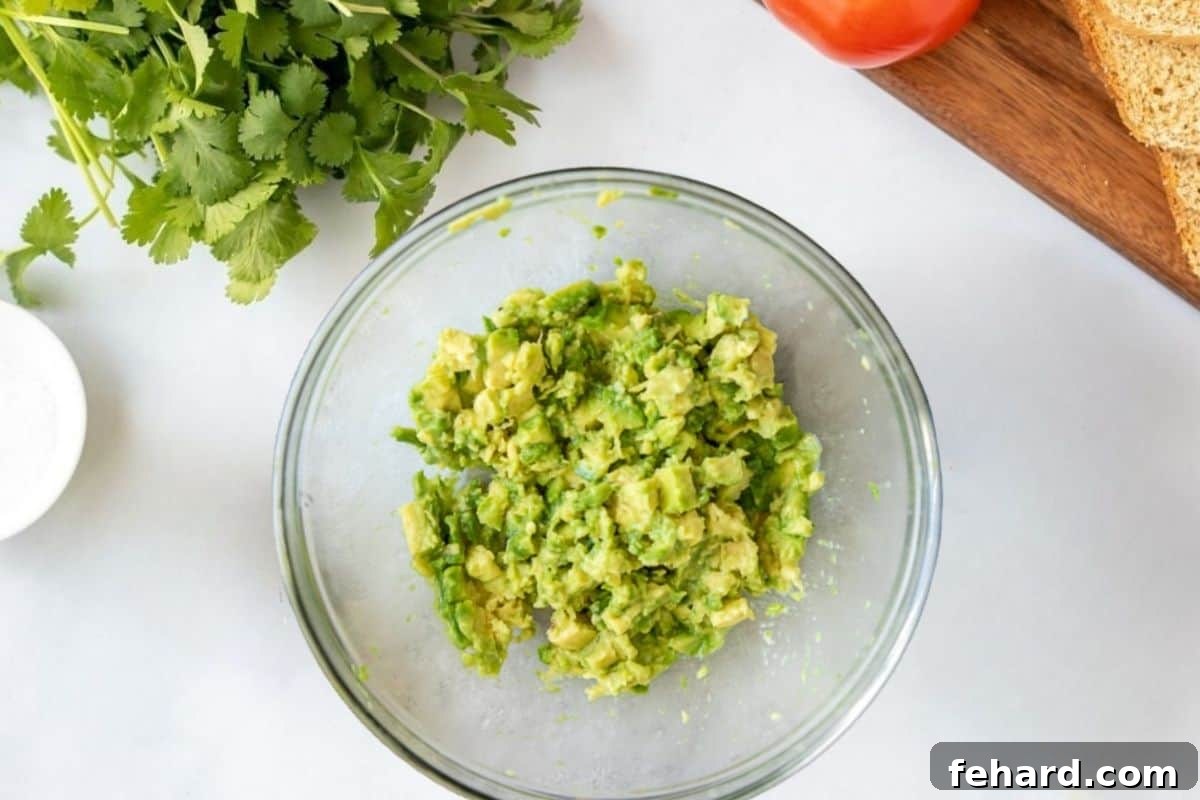 How to Mash Avocado for Chickpea Salad Mashed avocado with lime juice and seasonings in a large mixing bowl, ready for chickpeas to be added. Bread and tomatoes are visible on a cutting board nearby.