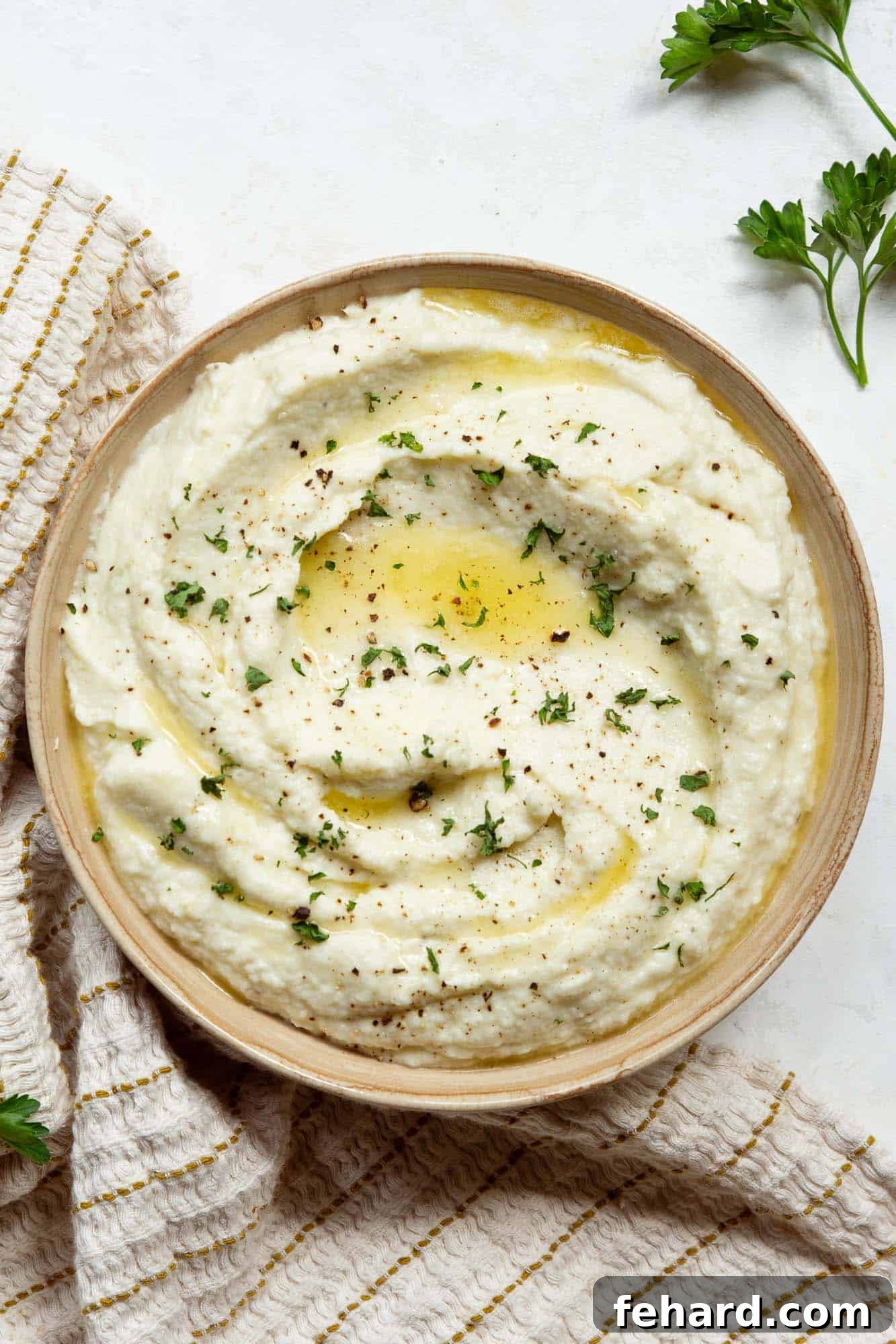 A bowl of mashed cauliflower on a marble countertop, garnished with fresh parsley.