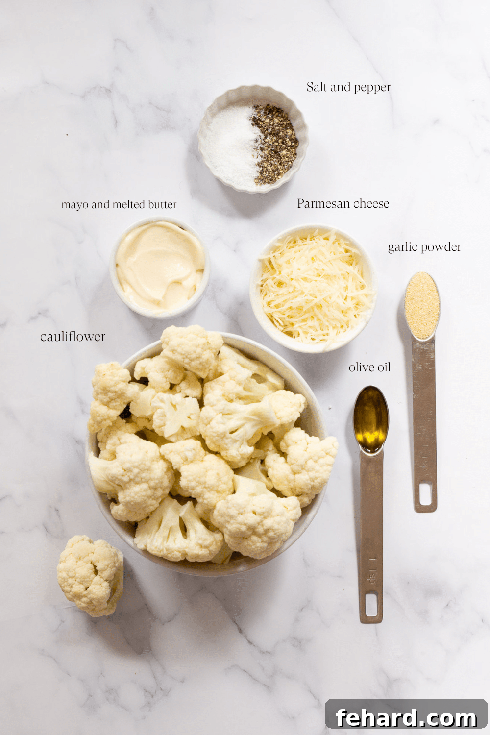 A collection of key ingredients for making mashed cauliflower on a marble countertop, including fresh cauliflower florets, a stick of butter, a jar of mayonnaise, and a wedge of Parmesan cheese.