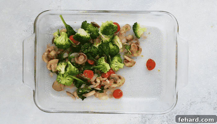 An overhead view of sautéed, colorful vegetables spread evenly in a square baking dish, forming the base of the breakfast casserole.