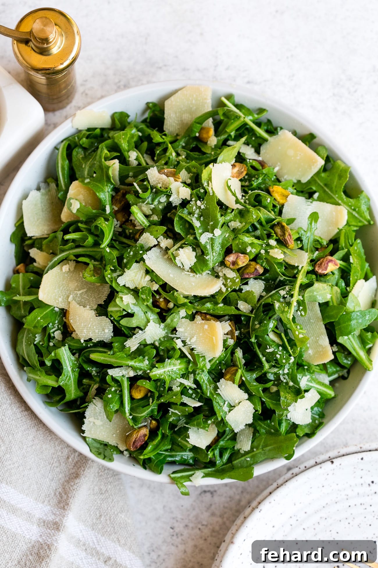 Overhead view of a fresh arugula salad in a white bowl, topped with Parmesan and pistachios.