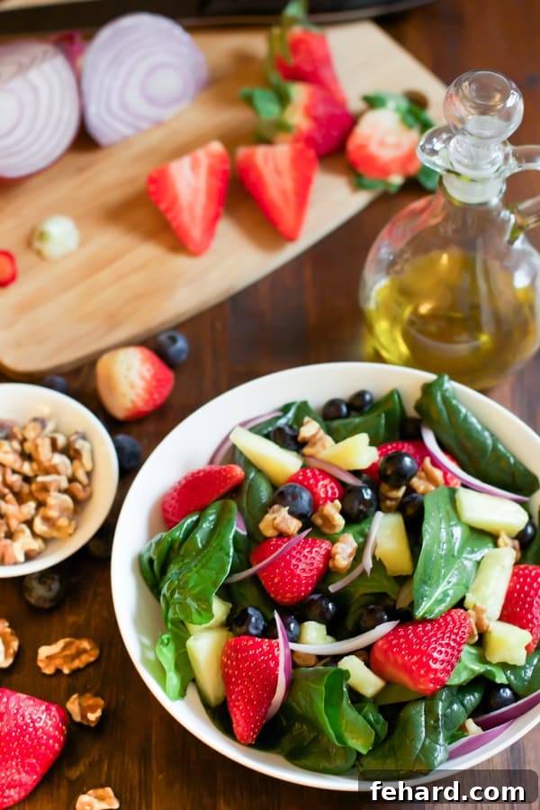 A bowl of fresh spinach fruit salad, brimming with colorful berries, pineapple, and walnuts, resting beside a cutting board with prepped ingredients.