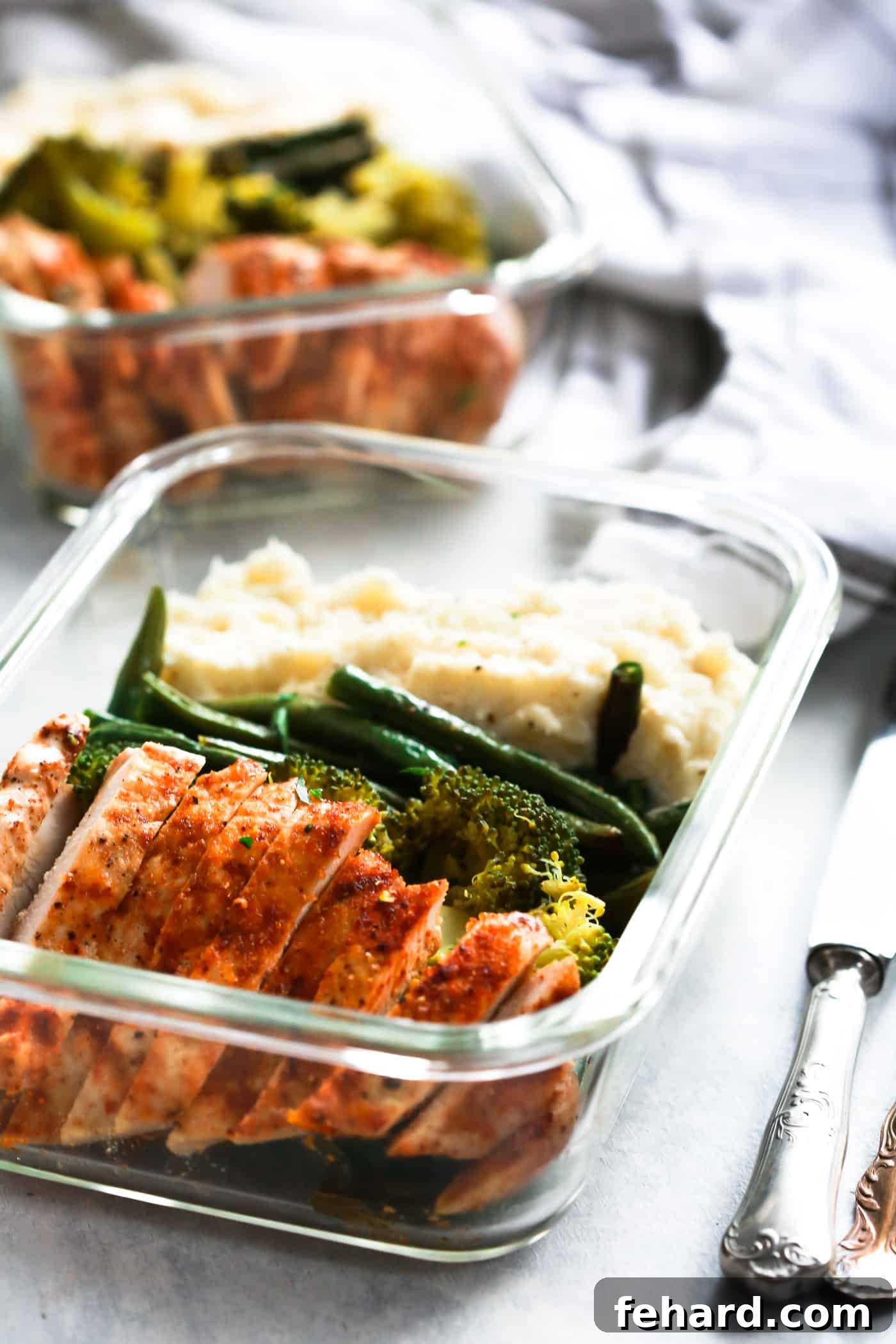 A clear glass container showcasing a well-balanced meal: spicy chicken, vibrant green broccoli, tender green beans, and a side of creamy mashed cauliflower.