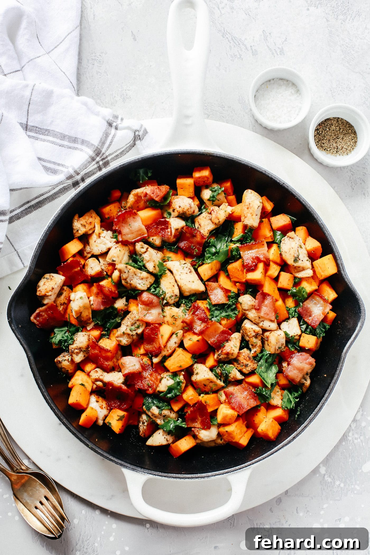 An overhead shot of a rustic cast-iron skillet, generously filled with tender chicken pieces, vibrant sweet potatoes, fresh kale, and crispy bacon.