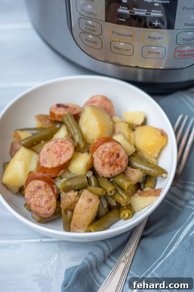 Instant Pot Smoked Sausage with Potatoes and Green Beans A white bowl filled with Instant Pot Smoked Sausage, Potatoes, and Green Beans, positioned next to an Instant Pot. The steam is visibly rising from the dish, emphasizing its freshness and warmth.