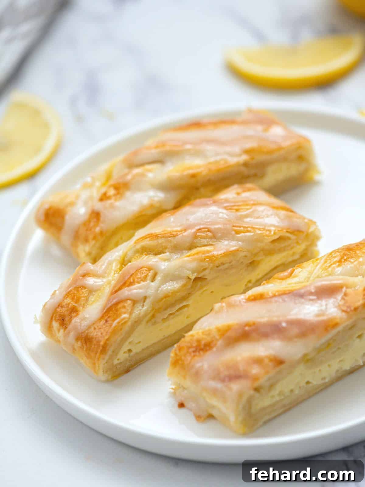 Three slices of a homemade cream cheese danish made with puff pastry and glazed with a lemon glaze on a white plate, showcasing its flaky layers and creamy filling.