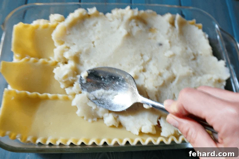 A hand spreading a layer of creamy mashed potatoes onto cooked lasagna noodles in a baking dish.