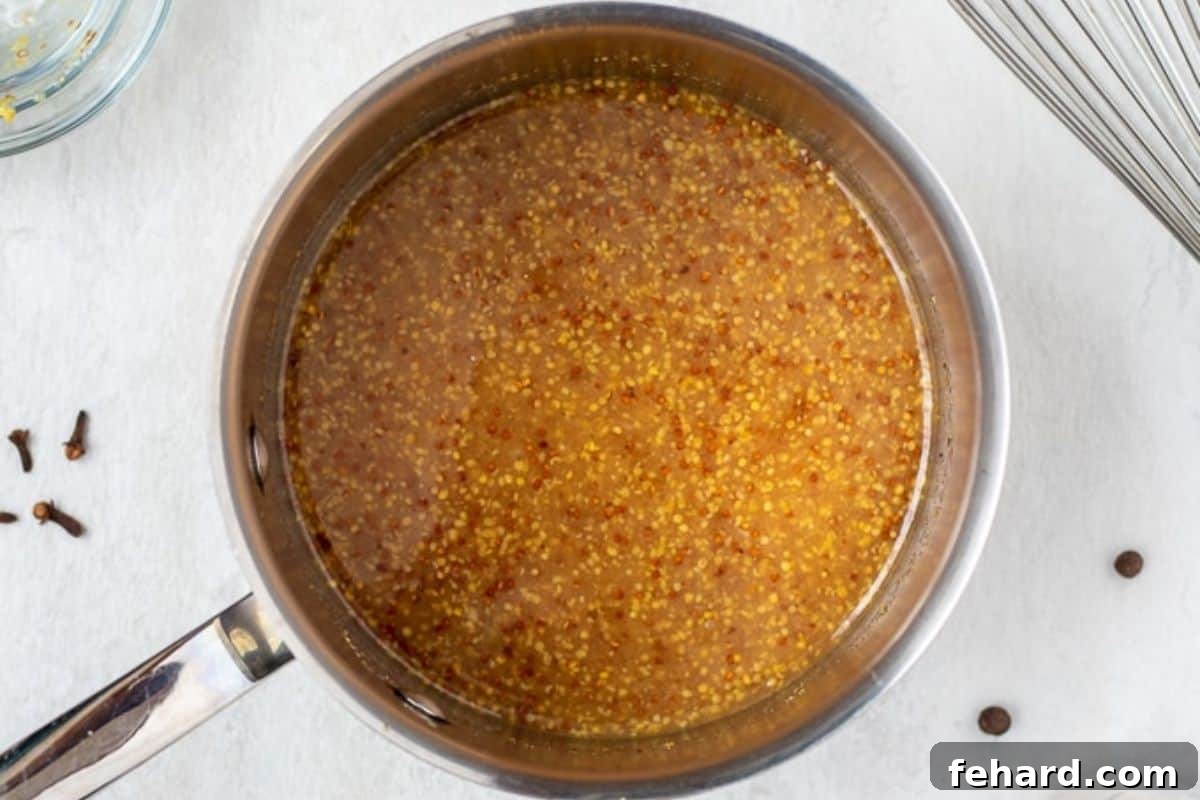 A small stockpot on the stovetop filled with a bubbling, golden pineapple Dijon mustard glaze, ready to be applied to the ham.