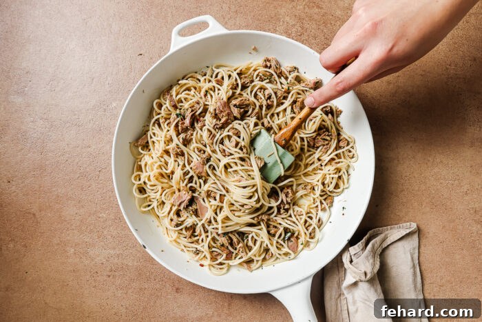 Cooked spaghetti being added directly to the skillet with the tuna and garlic sauce, ready to be tossed and combined.