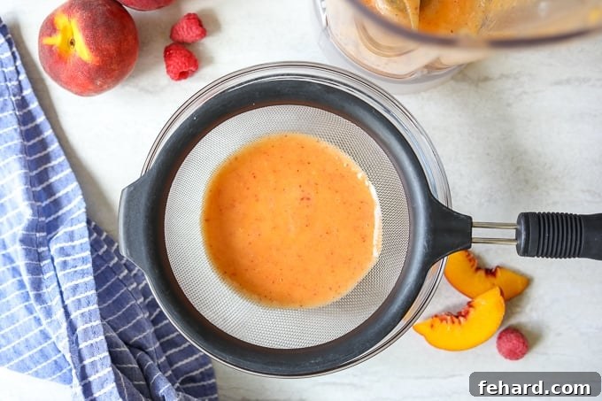 Freshly blended peach puree being strained through a fine-mesh strainer into a bowl, with a blender in the background, illustrating the crucial step for a smooth Bellini.