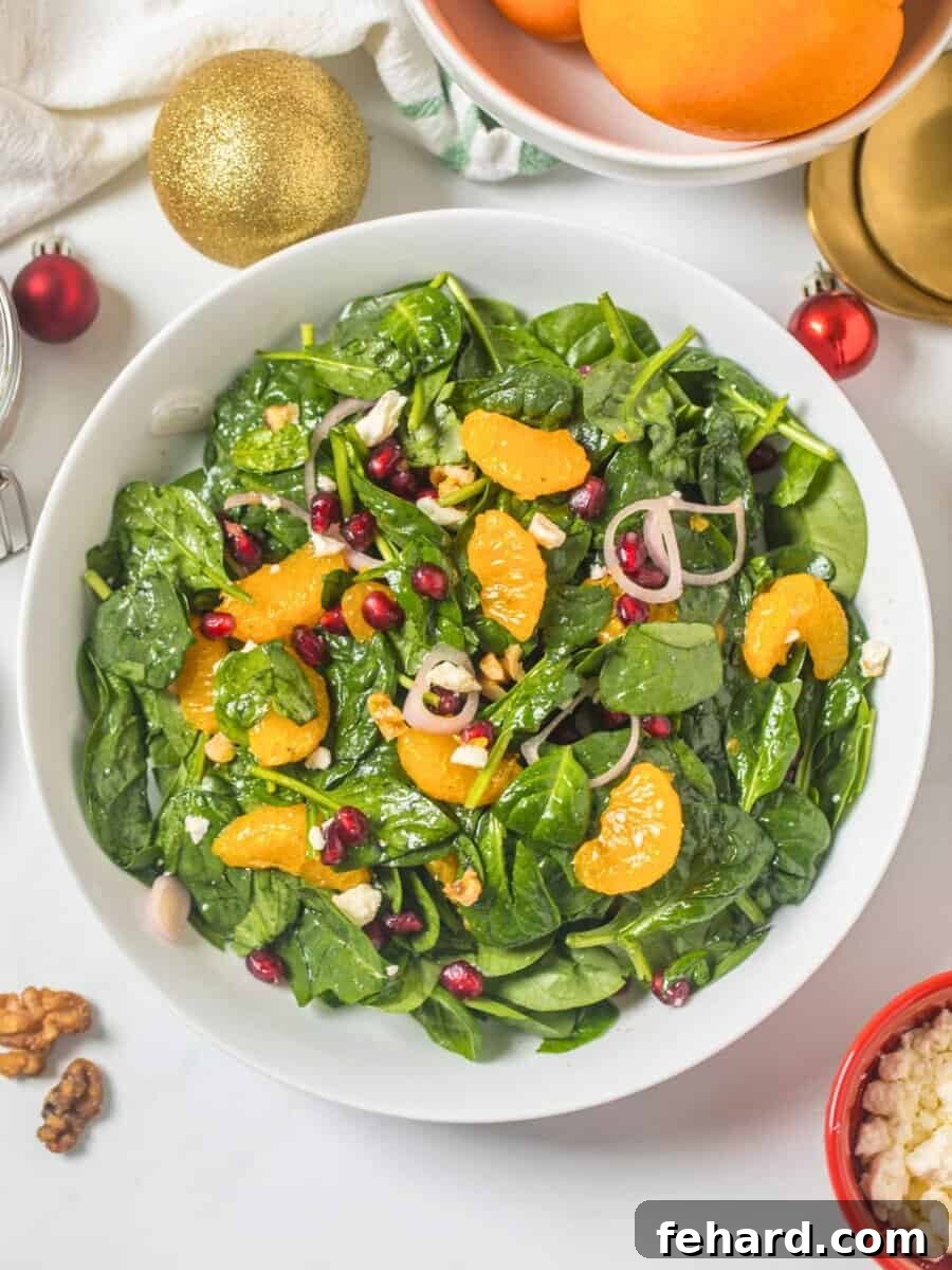 A beautifully arranged Christmas Salad in a white bowl, featuring baby spinach, vibrant mandarin orange segments, ruby red pomegranate arils, golden candied walnuts, crumbled white feta, and thinly sliced shallots, with a small bowl of champagne vinaigrette on the side.