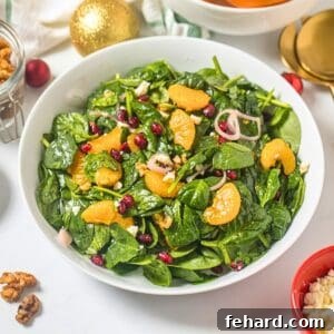 Salad made with baby spinach, mandarin oranges, pomegranate arils, candied walnuts, crumbled feta, shallots, and champagne vinaigrette in white bowl next to candied nuts and shallots.