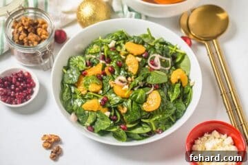 Salad made with baby spinach, mandarin oranges, pomegranate arils, candied walnuts, crumbled feta, shallots, and champagne vinaigrette in white bowl next to candied nuts and shallots.